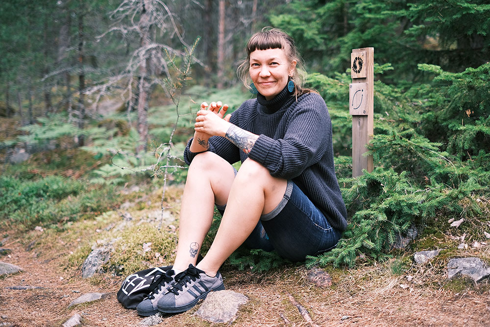 Jessica Koivistoinen sit down on the forest groudn by a sign on Barefoot path